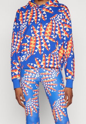 Personne portant un sweat à capuche bleu vif assorti et des leggings avec des motifs géométriques de feuilles rouges, jaunes et blanches ainsi que le logo Adidas sur la poitrine.