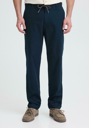 Mann trägt dunkelblaue, gerade geschnittene Hose mit Kordelzugbund und beigefarbene Wildleder-Schnürschuhe, steht vor einfachem Hintergrund.