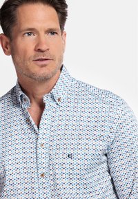 Chemise blanche à manches longues avec un motif abstrait bleu et marron, dotée d'un col et d'un logo discret sur la poche poitrine.
