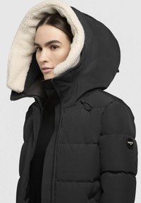 Frau trägt eine schwarze Daunenjacke mit einer großen Kapuze, die mit weißem Fleece gefüttert ist, und ist zugezippt. Dabei ist das Khujo-Markenbadge am Ärmel sichtbar.