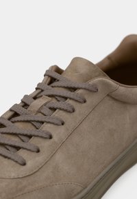 Taupe nubukbőr sneaker vastag, azonos színű fűzőkkel és gumitalppal, közelről, a felső elülső oldalra fókuszálva.
