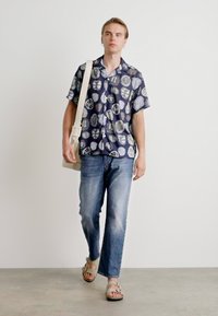 Camisa de manga curta, com botões, de fundo azul-marinho e padrões abstratos de cores claras, combinada com jeans azul claros e sandálias beges.