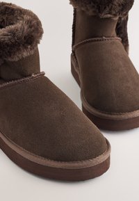 Hnědé semišové kotníkové boty s plyšovým kožešinovým límcem, zaoblenou špičkou a kontrastní hnědou gumovou podrážkou. Šité detaily podél okraje.