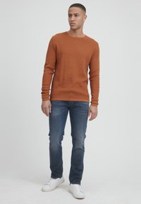 Pulover portocalie cu textură de vafe, cu mâneci lungi, asortat cu blugi albaștri și adidași albi, stând într-un fond alb simplu.