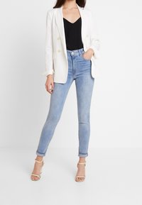 Blazer blanc à double boutonnage avec des boutons dorés, associé à un débardeur noir et un jean skinny bleu clair. Le mannequin porte des talons ouverts beige.