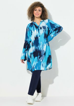 Lichtgewicht tuniek met lange mouwen in een blauw abstract patroon, gecombineerd met marineblauwe broeken. Heeft een kraag en een relaxte pasvorm. Draagt men met witte sneakers.