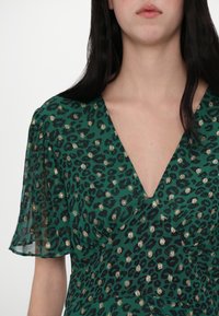 Groene jurk met luipaardprint, diepe V-hals, korte doorschijnende mouwen en metallic gouden accenten, gemaakt van een vloeiende, getextureerde stof.