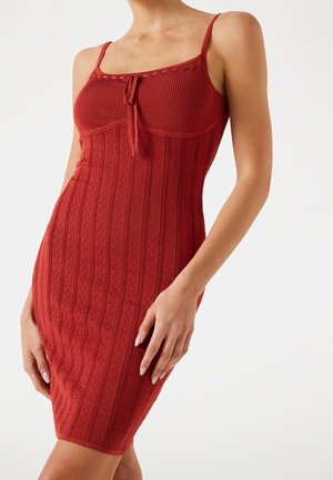 Frau trägt ein figurbetontes, ärmelloses, knielanges rotes Strickkleid mit dünnen Trägern und einer kleinen Schleifendetail am Ausschnitt.