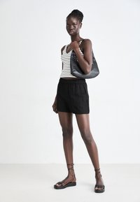 Débardeur rayé, shorts avec ceinture élastique noire, sac à main noir avec clous, et sandales noires à lanières. Le mannequin se tient devant un fond uni.