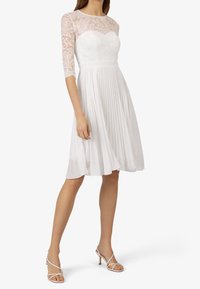 Robe blanche en dentelle et plissée, manches trois-quarts, corsage ajusté et jupe évasée. Associée à des talons blancs à brides.