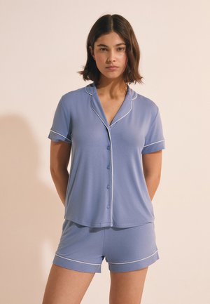 Femme aux cheveux courts et foncés portant un haut bleu clair à manches courtes avec boutons et un short assorti avec un passepoil blanc, debout devant un fond neutre.