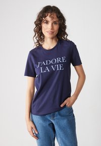 Námořnická modrá bavlněná tričko s nápisem "J'adore la vie" v světle modré. Krátké rukávy, kulatý výstřih, volný střih, v kombinaci s modrými džíny.