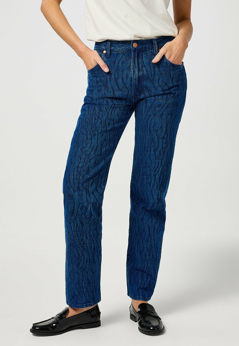 Jeans in denim blu con un motivo a onde, design a gamba dritta, stile classico a cinque tasche e vita con bottone; abbinati a scarpe nere lucide.