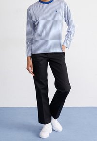 Långärmad randig skjorta i blått och vitt med blå krage, tillsammans med svarta byxor och vita sneakers, stående på ett blått golv.