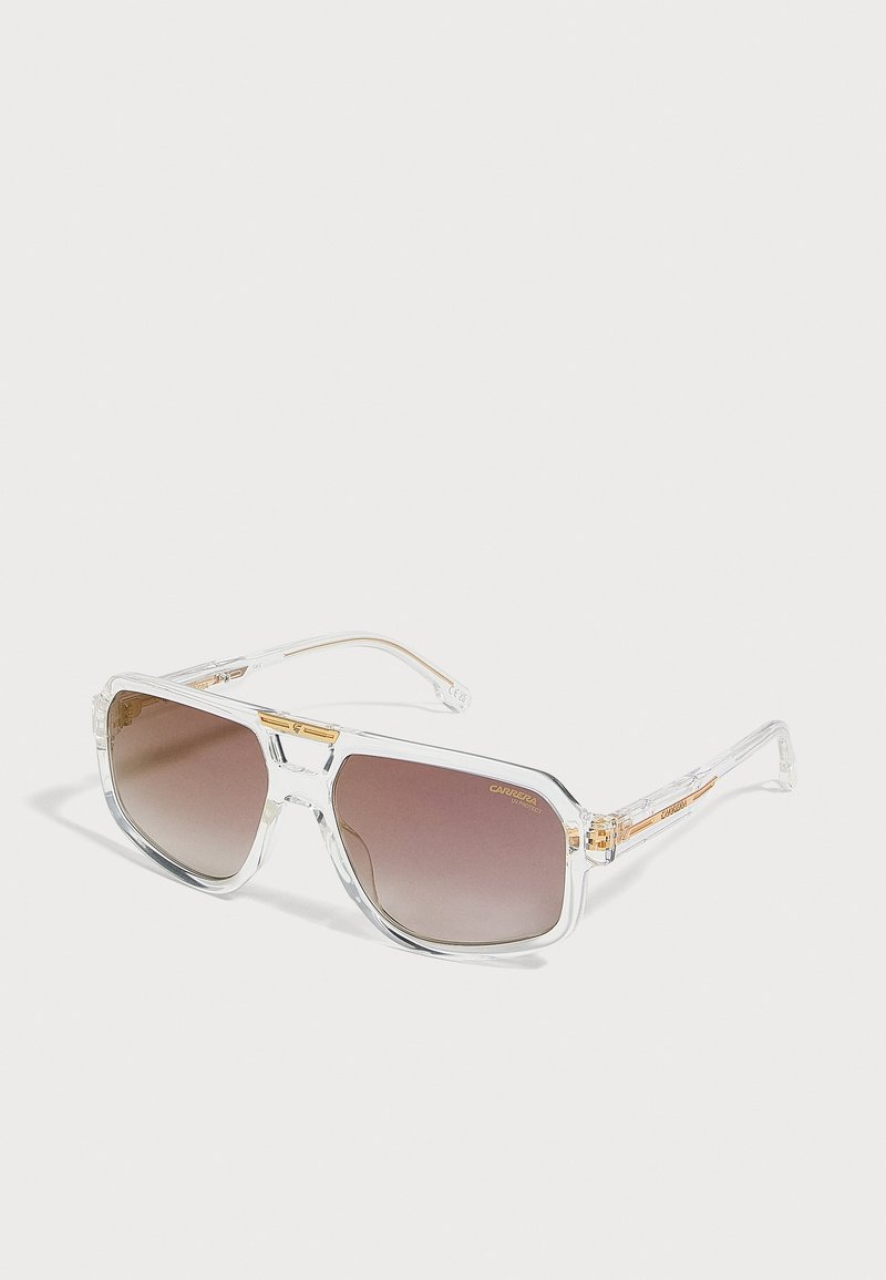 Lunettes de soleil carrées à monture transparente avec verres à dégradé marron et détails dorés sur les branches, reposant sur une surface claire.
