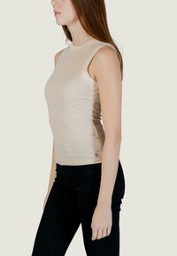 Frau mit langen Haaren, die ein ärmelloses beige Rippentop mit kleinen Glitzereffekten und eng anliegende schwarze Hose trägt, seitlich stehend.