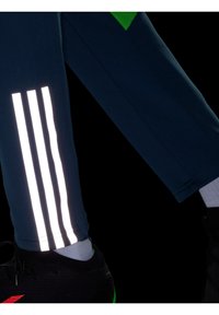 Marineblauwe sportbroek met drie reflecterende witte strepen aan de zijkant. Het materiaal lijkt glad, gecombineerd met zwarte schoenen met groene accenten.
