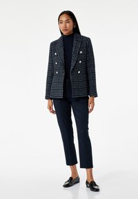 Vrouw in een marineblauwe geruiten dubbele breasted blazer, marineblauwe coltrui, slanke pantalons en zwarte instappers, staand tegen een witte achtergrond.