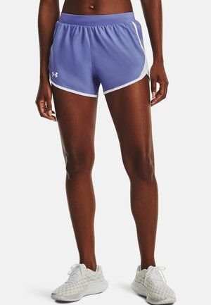 Athlète portant un short de course violet Under Armour avec bordure blanche et des chaussures de course blanches, debout devant un fond uni.