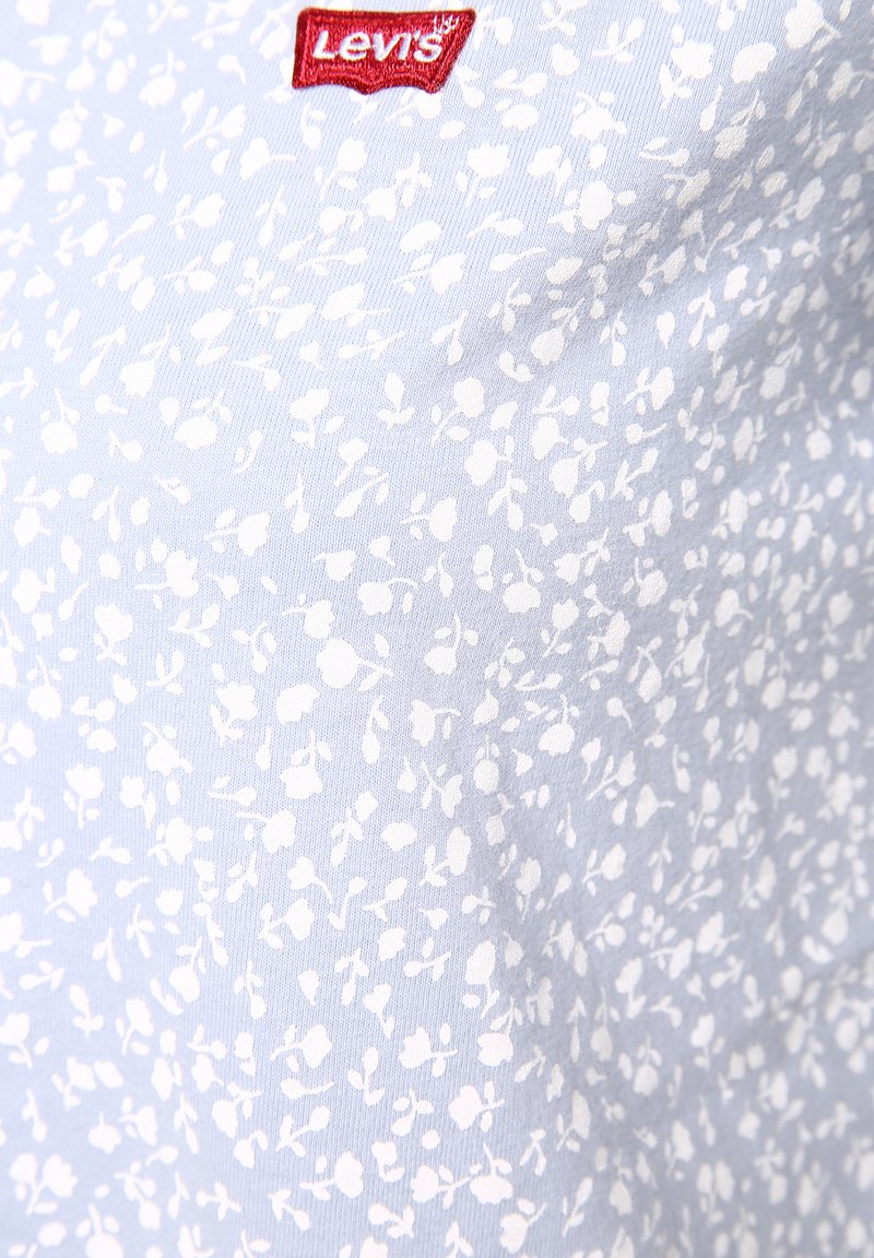 Tissu bleu clair avec un motif floral blanc ; comporte une étiquette logo rouge de Levi's. Texture douce, design ajusté, adapté à un style décontracté.