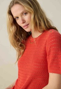 Pull court en tricot orange à manches courtes, présentant un motif texturé et des boutons décoratifs en métal doré à l'épaule. Design de col simple.
