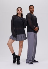 Due modelli indossano maglioni neri in maglia aperta; uno lo abbina a una gonna plissettata grigia e stivali neri, l'altro con pantaloni grigi e sneakers.