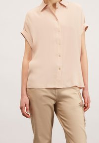 Světle růžová blůza s krátkým rukávem, límečkem, zapínáním na knoflíky vpředu a ohrnutými manžetami. Hladká textura s volným střihem.