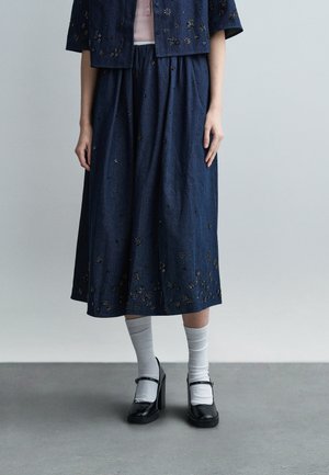 Frau trägt einen marineblauen Rock und eine Jacke mit Blumenstickerei, graue Socken und schwarze Mary Jane-Absatzschuhe, steht auf einem grauen Boden.