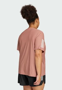 Maglietta sportiva color rosa con maniche corte, caratterizzata da tre strisce bianche sulla spalla sinistra e vestibilità morbida, abbinata a pantaloni corti neri.