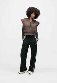 Jonge vrouw staat met handen in haar zakken, draagt een bruin mouwloos jack met rits en bontkraag, een zwarte top met lange mouwen, zwarte trainingsbroek en witte sneakers.