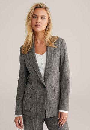 Grijze blazer met een houndstooth patroon, met geknipte revers, twee voorzakken en een enkele knoopsluiting, gelaagd over een witte top.