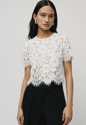 Paita renda putih dengan pola bunga, hem bergelombang, dan detail franja. Dipadukan dengan celana hitam high-waisted untuk kontras.