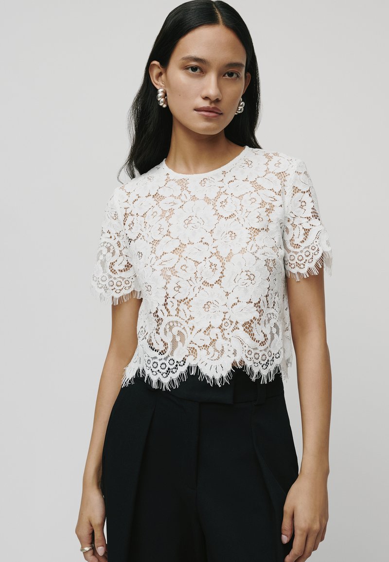 Witte kanten top met bloempatronen, gefronde zoom en franjes. Gecombineerd met zwarte high-waisted broeken voor contrast.