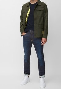 Chaqueta ligera de color verde oscuro con cuatro bolsillos frontales y un forro interior amarillo, combinada con vaqueros azules y zapatillas blancas.