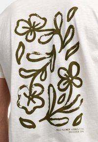 Hvid bomulds t-shirt med et mørkegrønt blomsterdesign på ryggen, med stiliserede blomster og blade samt tekst, der lyder "WILD FLOWER VIBES/1993."