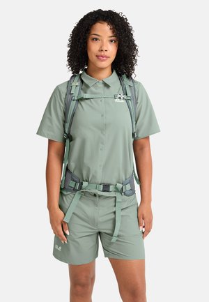Femme aux cheveux bouclés portant une chemise verte à manches courtes boutonnée, un short assorti, et un sac à dos avec des sangles poitrine et taille.