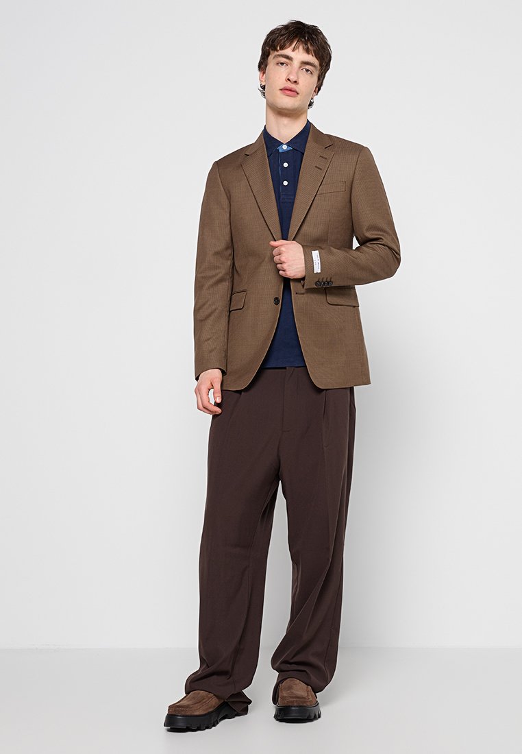 Junger Mann steht, trägt einen braunen Blazer, ein dunkelblaues Polohemd, lockere dunkelbraune Hose und braune Schuhe vor einem einfarbigen hellen Hintergrund.