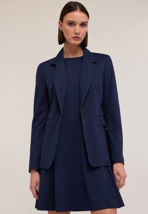 Marineblazer met geknipte revers, enkele knoopsluiting en twee zijzakken, gecombineerd met een bijpassende marineblauwe jurk. Soepele stoftextuur.