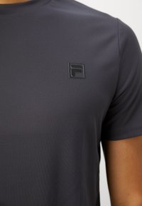 Nærbillede af en person, der bærer en mørkegrå t-shirt med korte ærmer og et lille sort Fila-logo på brystet.