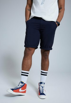 Dunkelblaue Shorts mit umgeschlagenem Saum, kombiniert mit hochgeschnittenen orange-blauen Sneakers und weißen Socken mit schwarzen Streifen.