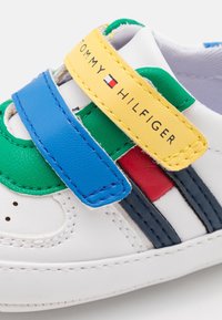 Białe sneakersy z niebieskimi, zielonymi, czerwonymi i granatowymi akcentami. Posiadają żółty pasek z napisem "TOMMY HILFIGER" oraz gładką skórzaną teksturę.