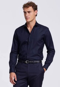 Marineblauwe button-up shirt met lange mouwen, slim fit, gladde stof en subtiele glans. Draagt met een zwarte leren riem.