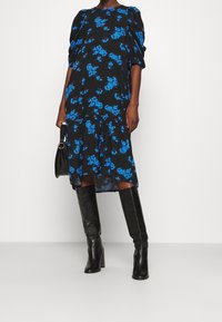 Černé květinové šaty s modrými vzory, krátké nabírané rukávy, pružný pas a splývavý lem. Nosené s černými kozačkami po kolena a malou kabelkou.