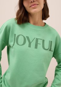 Sudadera verde claro con el texto "ALEGRÍA" en un verde más oscuro. Cuenta con un cuello redondo y una textura suave y lisa.
