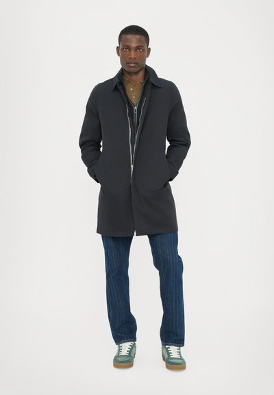 Donkerblauwe jas met een gladde textuur, rits sluiting en zijzakken, gecombineerd met blauw gestreepte jeans en groene sneakers met witte accenten.