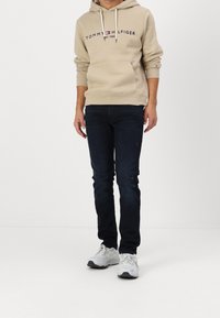 Beige cotton hoodie with front pocket and logo, paired with dark blue jeans featuring slight distressing and white sneakers.