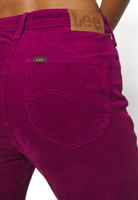 Gros plan d'un pantalon en velours côtelé magenta vif avec poche arrière et étiquette de marque Lee en cuir à la taille.