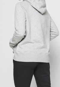 Felpa grigia con una texture morbida, dotata di cappuccio con cordino, polsini a coste e tasche frontali. Indossata con pantaloni neri.