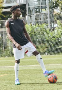 Svart ärmlös idrottströja, vita shorts med röda accenter, vita strumpor och flerfärgade fotbollsskor, dribblar en svart och orange fotboll.
