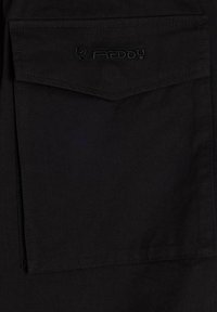 Camisa de tela negra con un bolsillo en el pecho que presenta un detalle de logo bordado. El material es suave, con un acabado mate y líneas limpias.